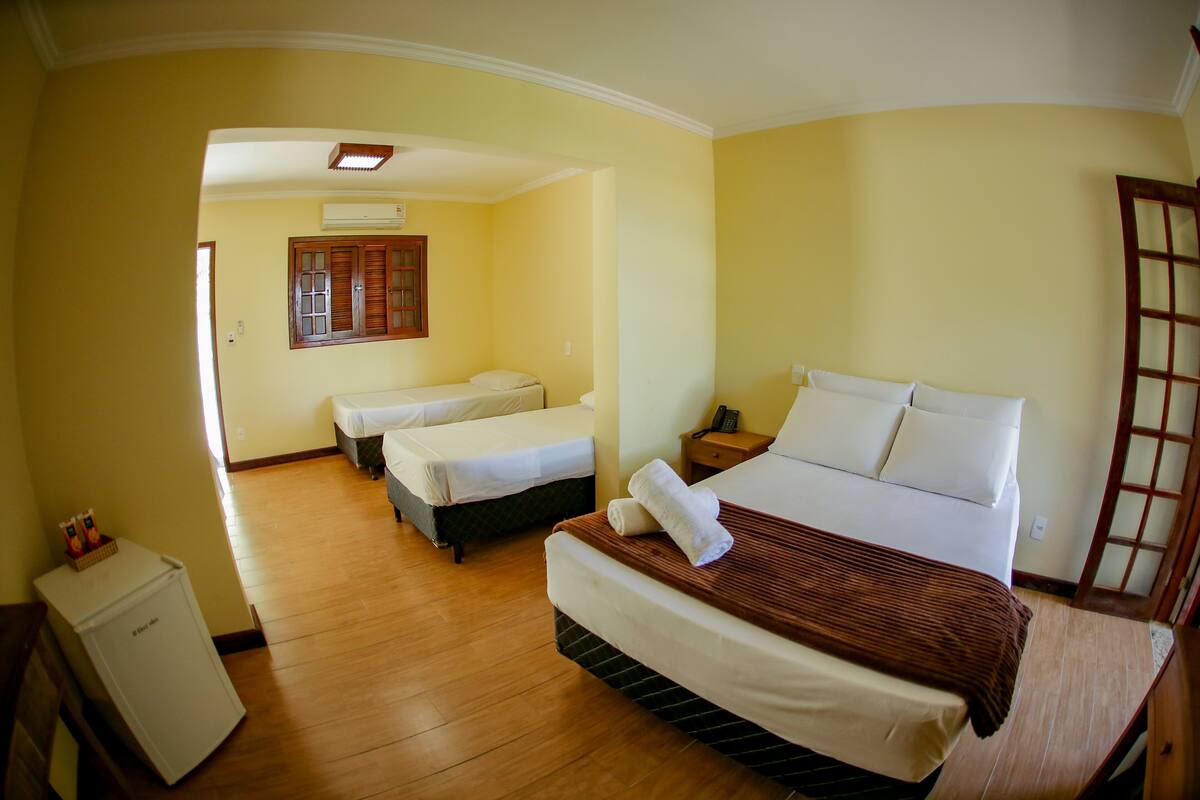 A hotel room with two single beds, a large bed, wooden flooring, a small refrigerator, and a wooden wardrobe.