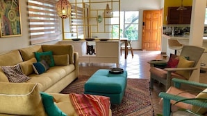 Uma sala de estar com sofá, puf e poltronas, área de cozinha com mesa de jantar e uma grande janela com persianas.