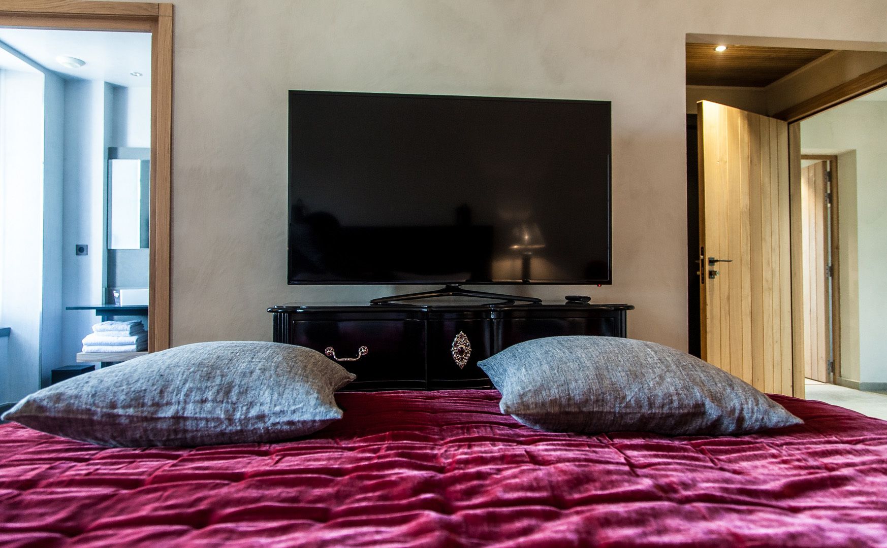 Un dormitorio con una televisión de pantalla plana grande montada en la pared, una cama con una colcha roja y dos almohadas.