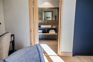 Une chambre d’hôtel avec une porte à cadre en bois donnant sur une salle de bain équipée d’un miroir, d’un lavabo et d’un espace pour ranger les serviettes.
