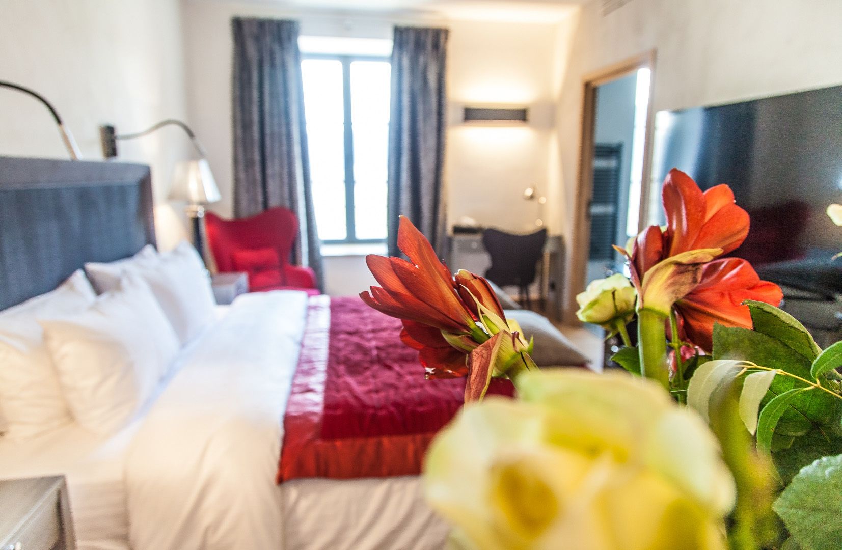 Habitación de hotel con una cama, un escritorio y una silla. Hay flores sobre la cama y una ventana con cortinas.