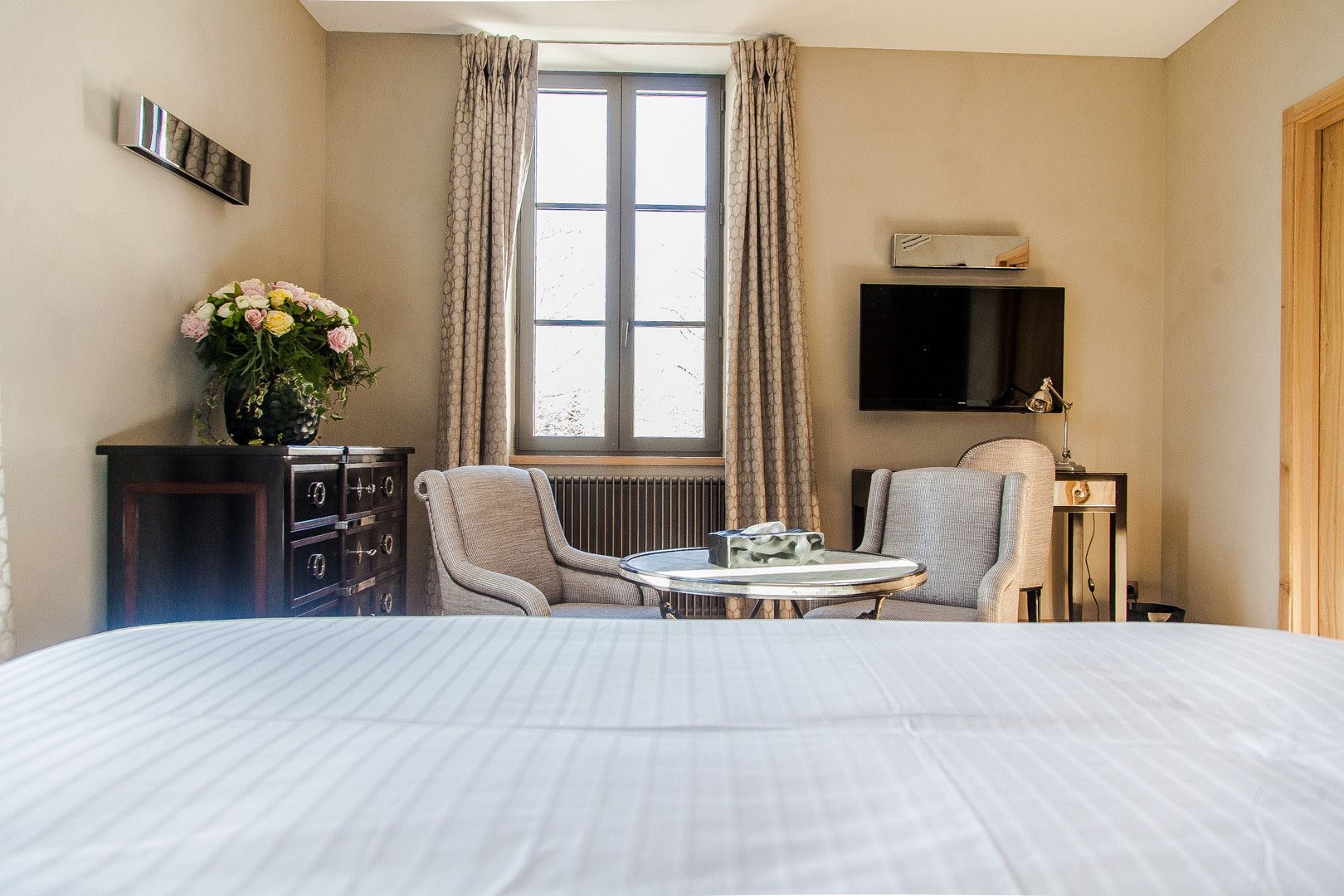 A hotel room with a large bed, two armchairs, a small table, a dresser, a TV, and a window with curtains.