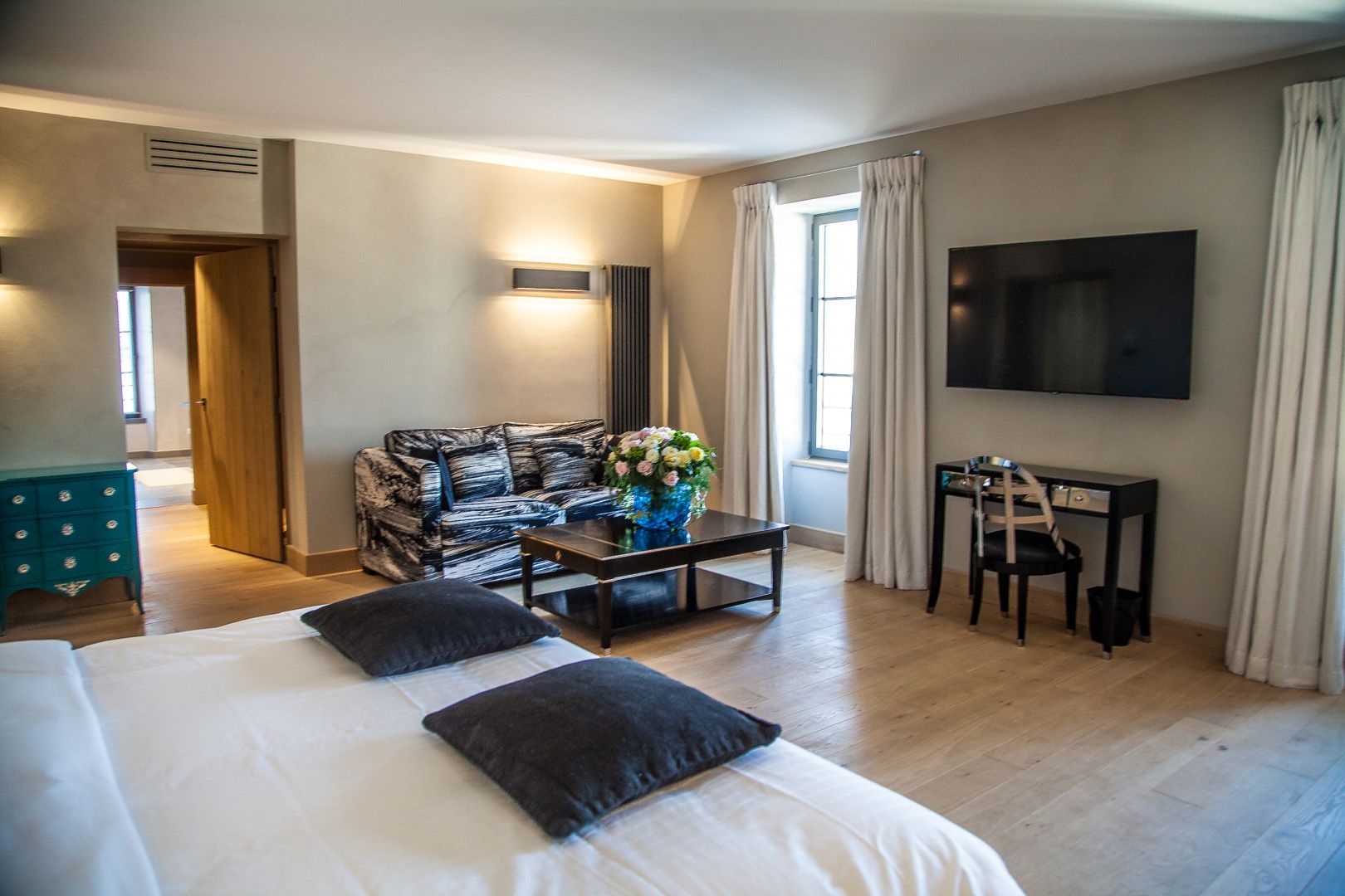Habitación de hotel con una cama grande, un sofá, una mesa de centro con flores, un escritorio con una silla y un televisor en la pared.