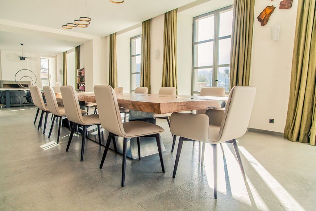 Een ruime eethoek met een grote houten tafel en meerdere stoelen, grote ramen met gordijnen, en een moderne plafondlamp.