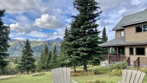 A patio with two Adirondack chairs and a fire pit, overlooking a mountainous landscape with a house in the background. - Impressive Mountain Views. Craftsman Home.  Less than 10 miles to bus depot (denali)