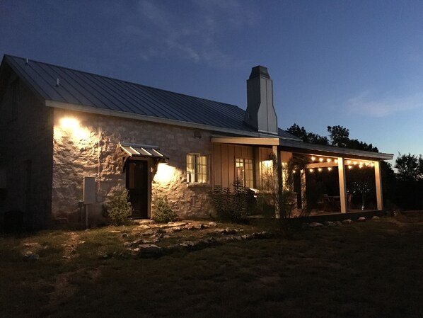 A stone house with a metal roof, a chimney, and a porch with string lights. - Heaven in the Hills. Welcome to Monticello Cottage (Dripping Springs)