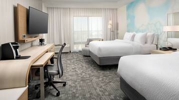 A hotel room with a flat-screen TV mounted on the wall, a desk with a chair, a bed with white bedding, and a large window with curtains.