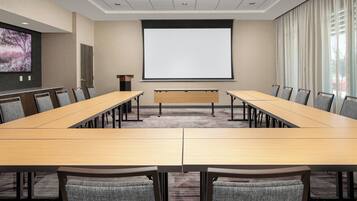 Ein Konferenzraum mit einem langen Tisch, mehreren Stühlen, einer Projektionsleinwand und einem großen Fenster mit Vorhängen.
