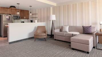 A modern living room with a sofa, armchair, and a small table. There is a kitchen area with a refrigerator, microwave, and cabinets.