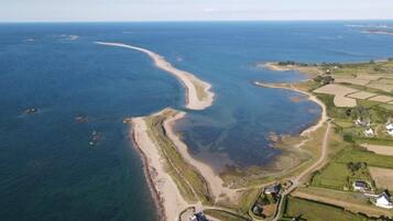 An aerial view of a coastal area with a sandy beach, clear blue waters, and residential properties with houses and fields.