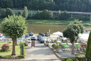 Un área de estacionamiento junto al río, con coches, un café con terraza y un puente al fondo.