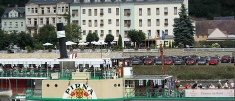 A riverboat with 'PIRNA' written on its side, docked near a large hotel with multiple stories and balconies.