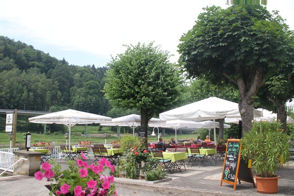 Un área de comedor al aire libre con mesas y sombrillas, rodeada de follaje y árboles.
