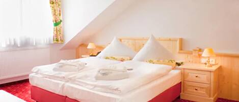 A hotel room with a red bed, white pillows, a wooden headboard, a nightstand with a lamp, and a window with curtains.