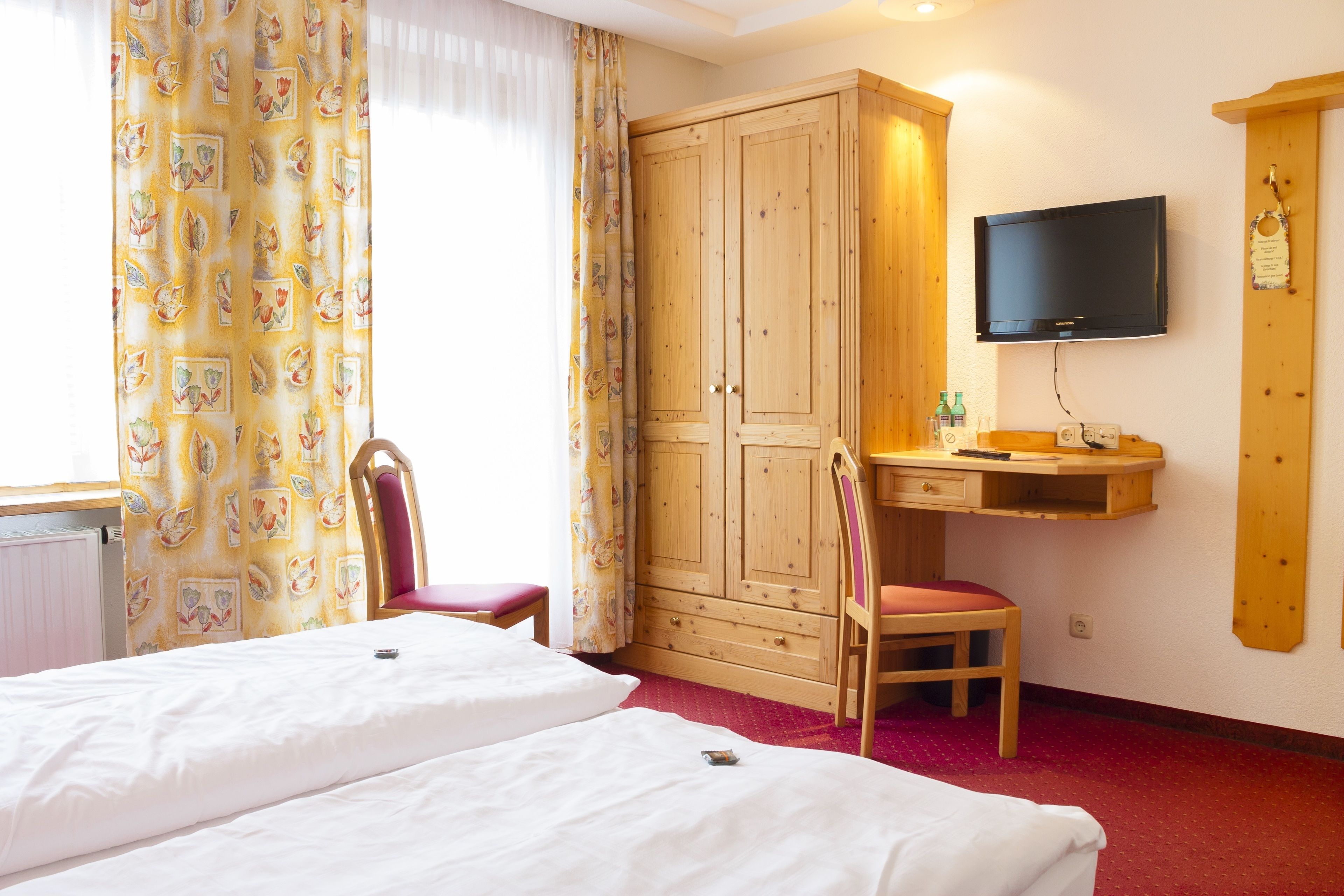 A hotel room with two beds, a wooden wardrobe, a desk with a chair, a television, and a red carpet.