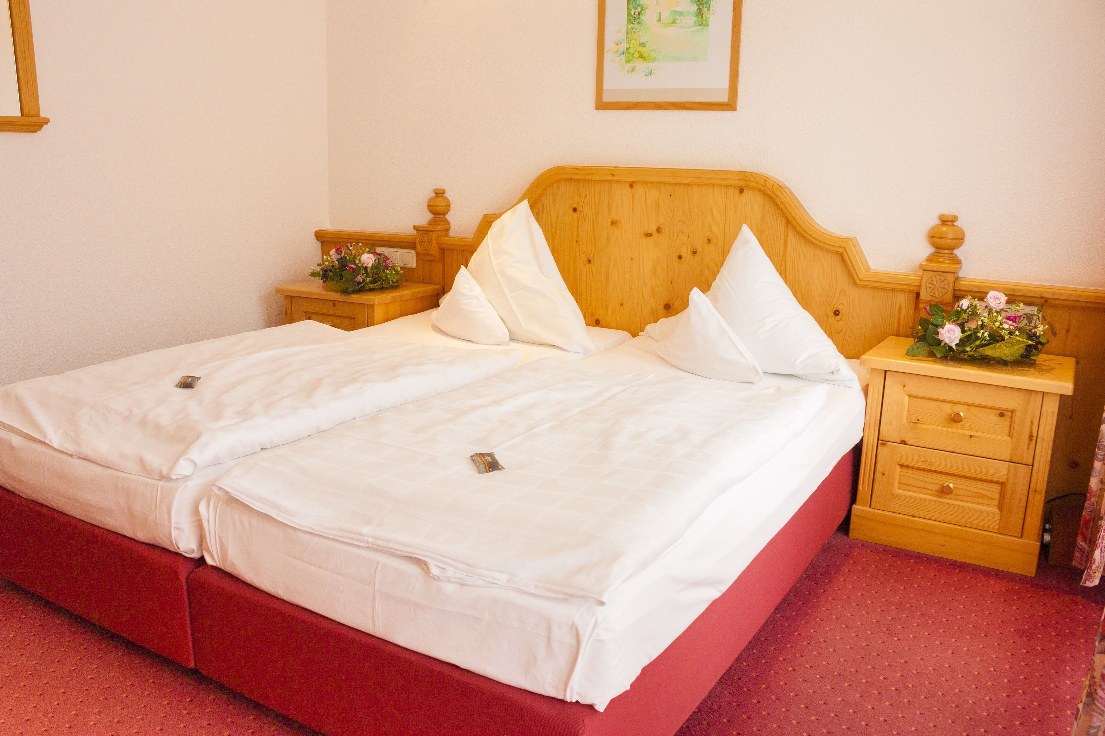 A double bed with white linens, a wooden headboard, and a red accent on the bed frame. There are two bedside tables with lamps and a small bouquet of flowers on one.