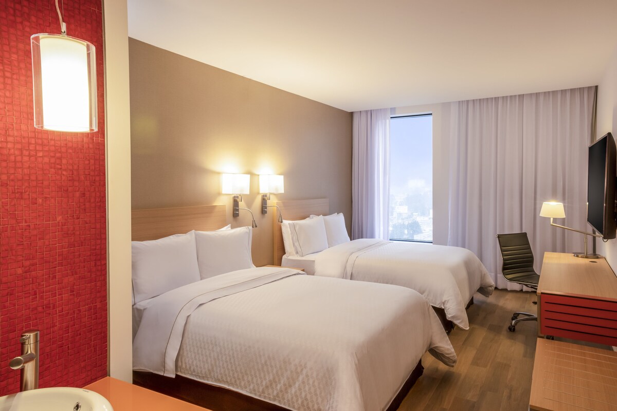 A hotel room with two beds, a desk, a TV, and a window with curtains.
