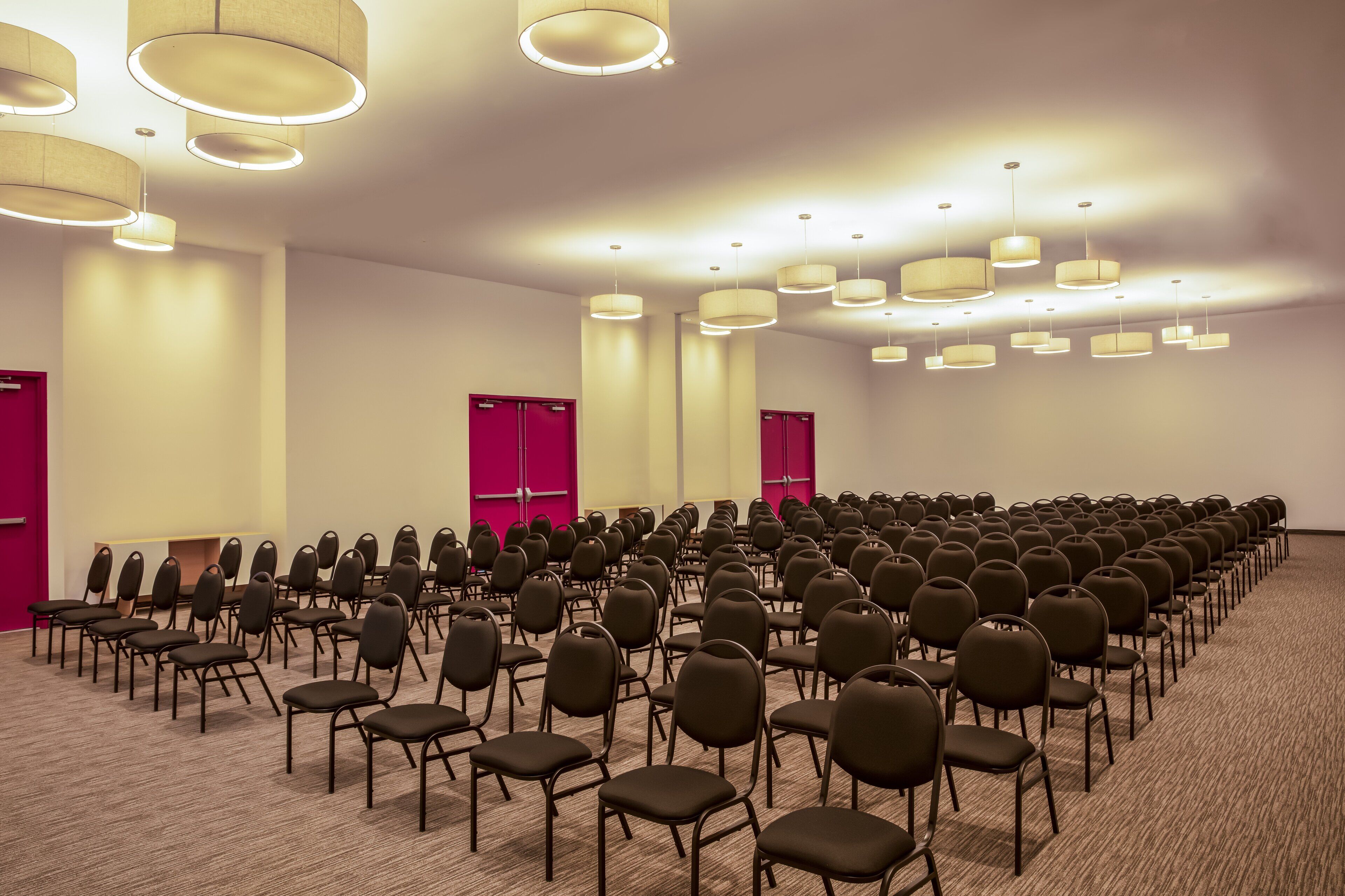 Un salón de conferencias con filas de sillas, luces en el techo y puertas rojas.