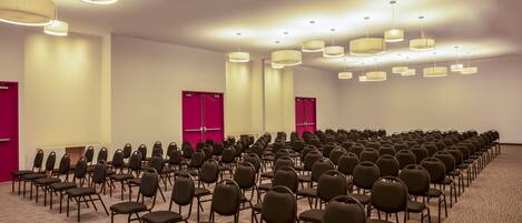 Un salón de conferencias con filas de sillas, luces en el techo y puertas rojas.