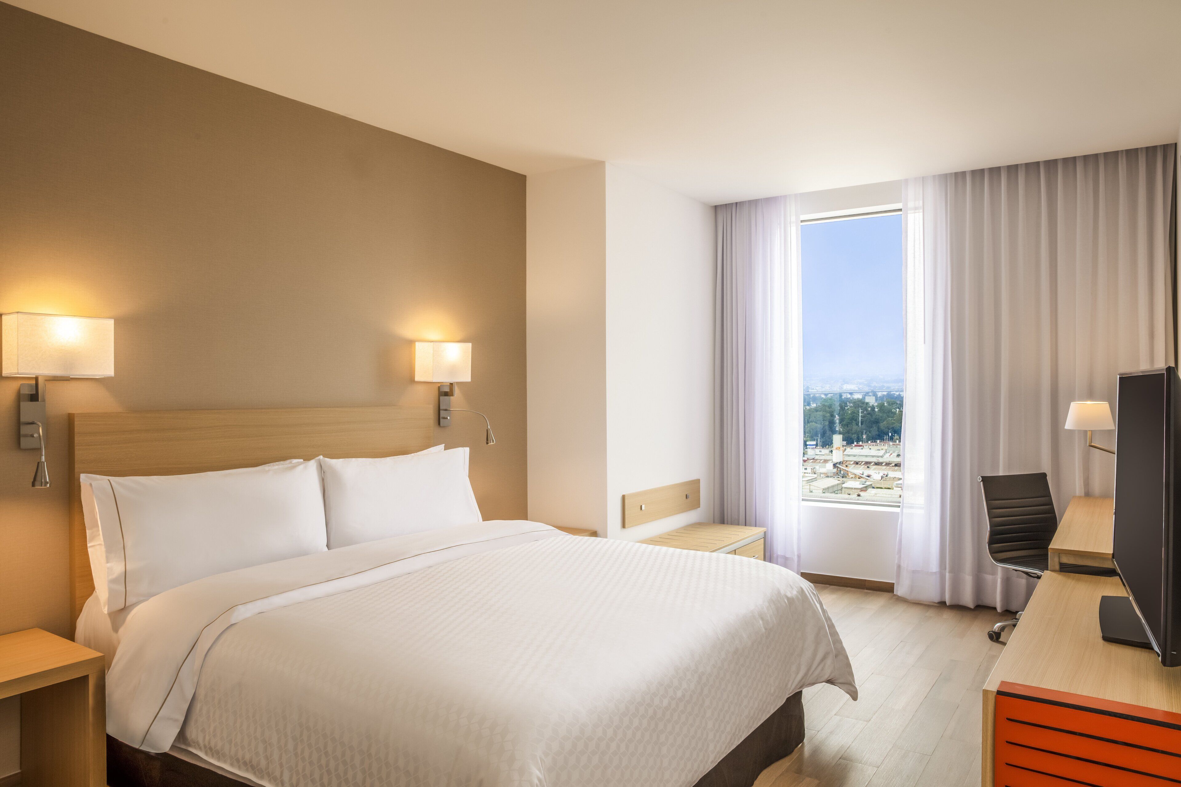 Habitación de hotel con una cama grande, dos lámparas de noche, un escritorio con silla, televisor y ventana con cortinas.