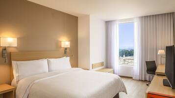 Habitación de hotel con una cama grande, dos lámparas de noche, un escritorio con silla, televisor y ventana con cortinas.