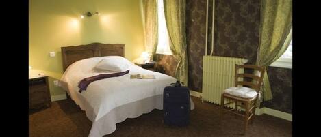 A bedroom with a bed, a chair, a nightstand, a lamp, a suitcase, and a radiator.
