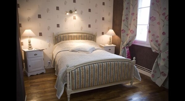 A bedroom with a bed, two bedside tables, a nightstand, a window with curtains, and a wall with a floral pattern. - Le Lion dOr (Écouché-les-Vallées)