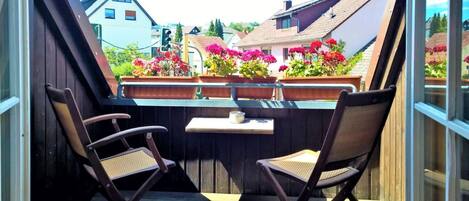 A balcony with two chairs and a small table, overlooking a row of colorful flowers and houses.