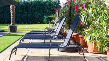A row of black lounge chairs under a canopy, with potted plants and a neatly trimmed lawn in the background.