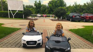 Zwei Kinder fahren Spielzeugautos auf einer befestigten Fläche mit kreisförmigem Muster, umgeben von Grünpflanzen und geparkten Autos.