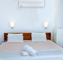 Una habitación de hotel con cabecera de madera, dos luces fijadas a la pared, una cama con colcha estampada en rosa y un aire acondicionado sobre la cama.