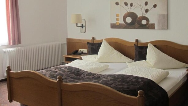 Ein Hotelzimmer mit einem hölzernen Bett, einem Nachttisch mit Lampe, einem Heizkörper und einem Bild an der Wand.