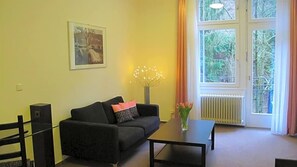 A living room with a sofa, a coffee table, a picture on the wall, a vase with flowers, and a window with curtains.