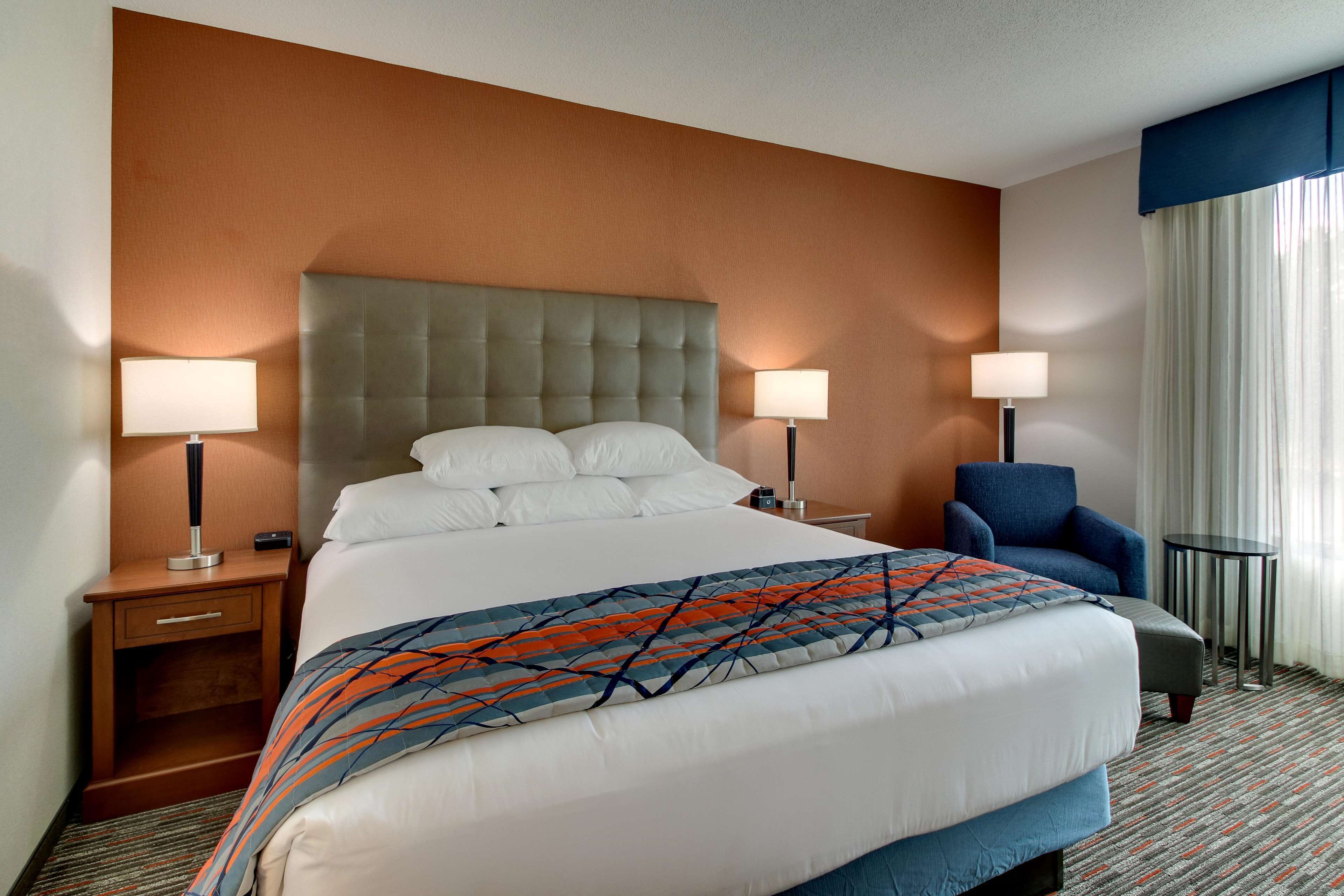 A hotel room with a large bed, two bedside tables with lamps, a chair, and a view of the outdoors through a window with curtains.