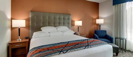 A hotel room with a large bed, two bedside tables with lamps, a chair, and a view of the outdoors through a window with curtains.