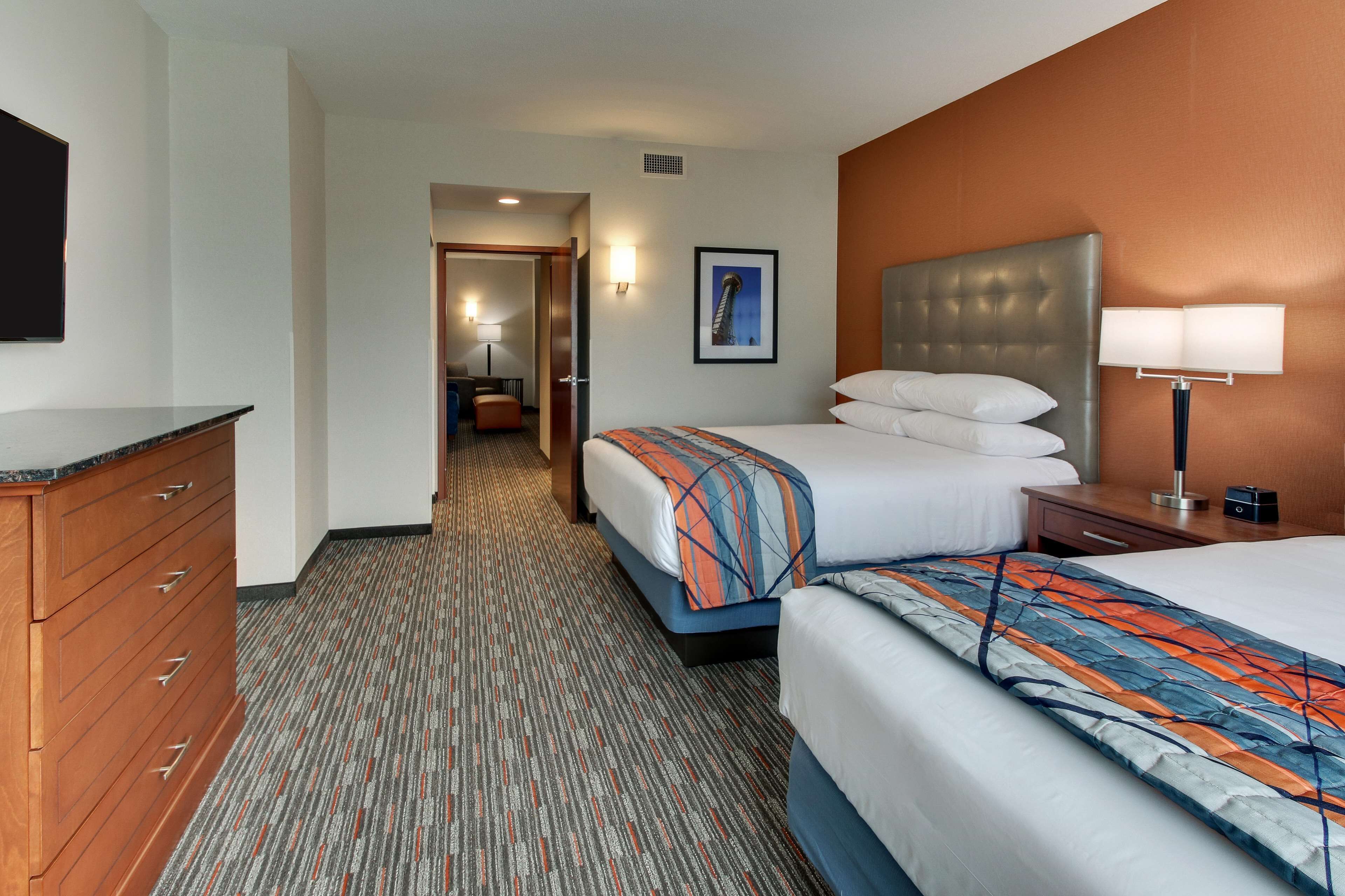 A hotel room with two beds, a wooden dresser, a desk with a lamp, and a framed picture on the wall.