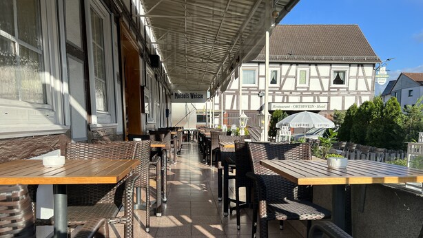 An outdoor seating area with wicker chairs and wooden tables, a covered walkway, and a building with a sign for 'ORTWEIN-Hotel' in the background. - Hotel Orthwein (Coelbe)