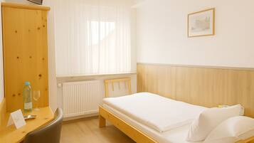 Une chambre d’hôtel avec un lit en bois, un petit bureau avec une chaise, un radiateur et une fenêtre avec des rideaux légers.