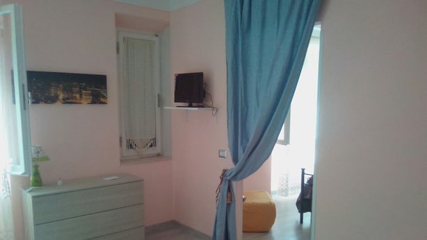 A bedroom with a dresser, a bed, a window with curtains, and a small shelf with a television. - Sweet Dream (Passignano sul Trasimeno)