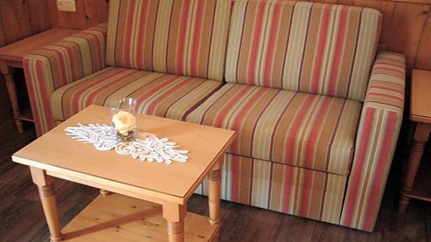A wooden sofa with striped upholstery, a small wooden table with a doily and a vase, and a wooden floor. - Alpenhotel Pfaffenwinkel (Peiting)
