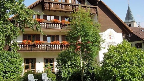 A multi-story building with balconies, a sign indicating it's an 'Alpenhotel,' and a church spire in the background.