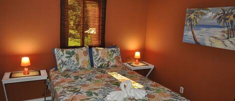 A bedroom with a floral bedspread, two bedside lamps, a painting of a beach scene, and a window with blinds.