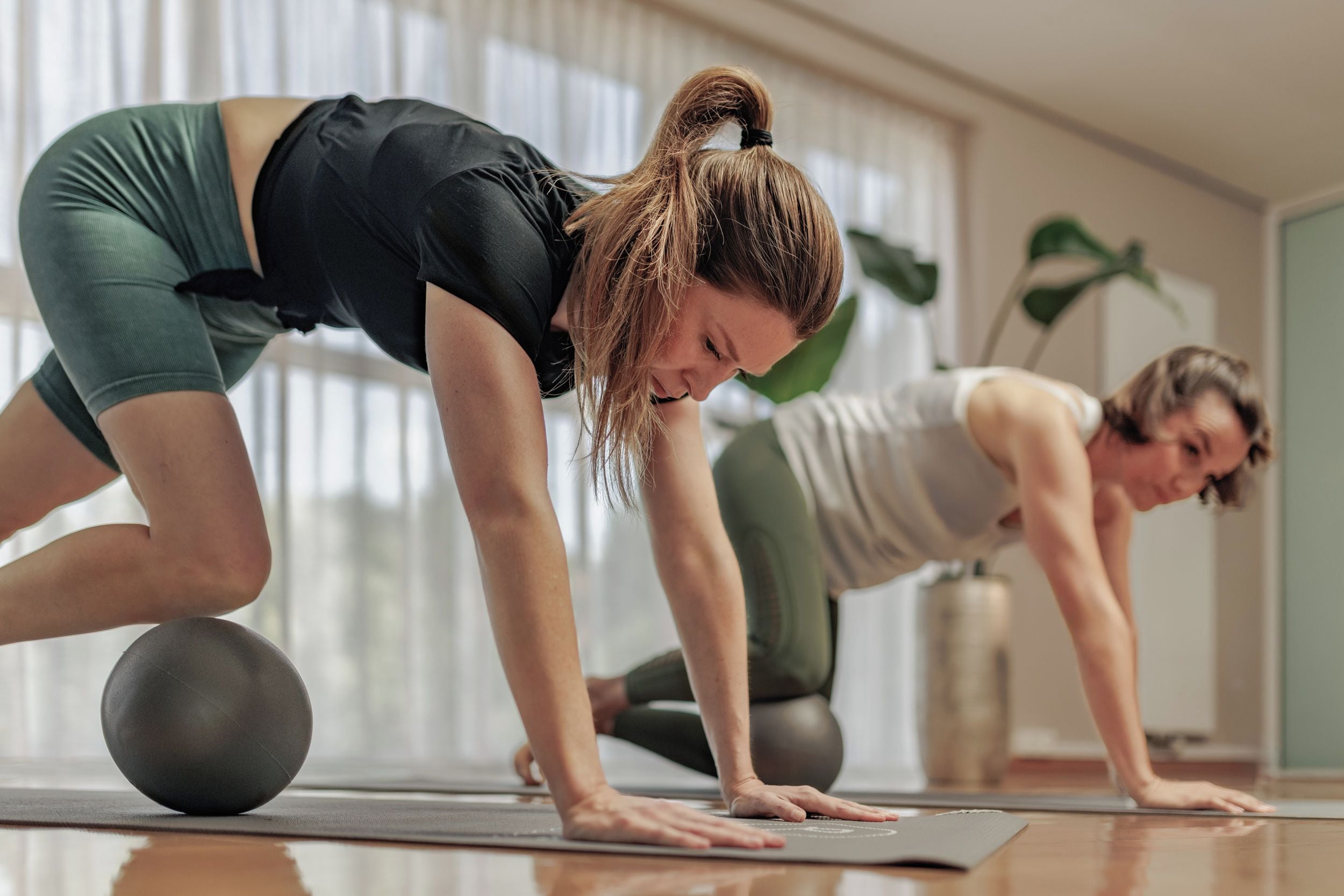 Deux personnes pratiquent une posture de yoga sur des tapis de sport dans une pièce où se trouvent une grande plante et une fenêtre.