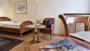 A hotel room with a bed, a small round table with a lamp, a chair, and a desk with a laptop and magazines.