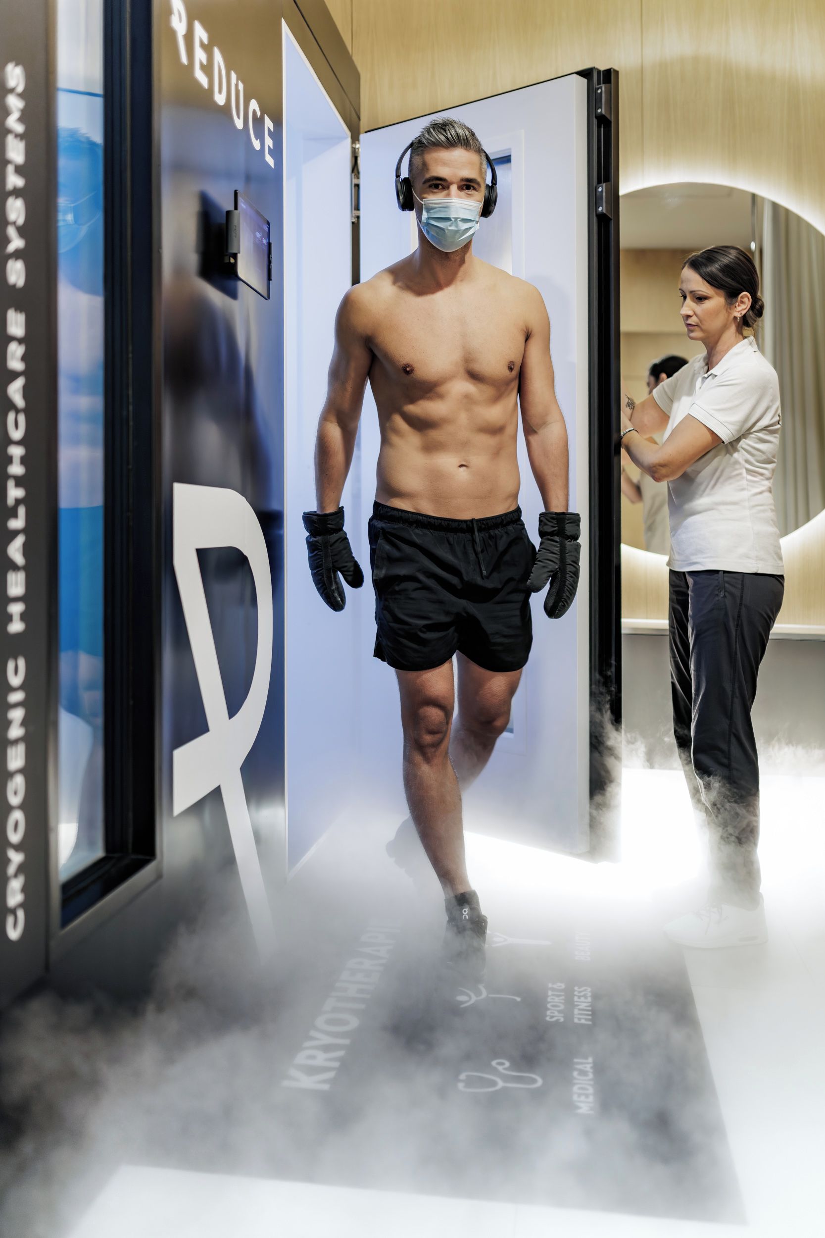 Un hombre camina por una puerta con una gran letra 'R' en la pared, un cartel que dice 'CRYOGENIC HEALTHCARE SYSTEM', y una persona al fondo sosteniendo un micrófono.