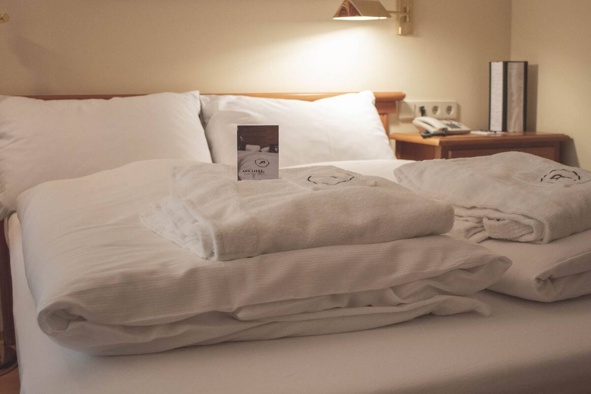 Ein ordentlich bezogenes Bett mit weißen Leinen und Handtüchern, ein Nachttisch mit Lampe und ein gerahmtes Bild an der Wand.