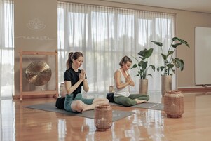 Dos personas sentadas sobre esterillas de yoga en una habitación con ventanas amplias, suelo de madera y plantas en macetas.