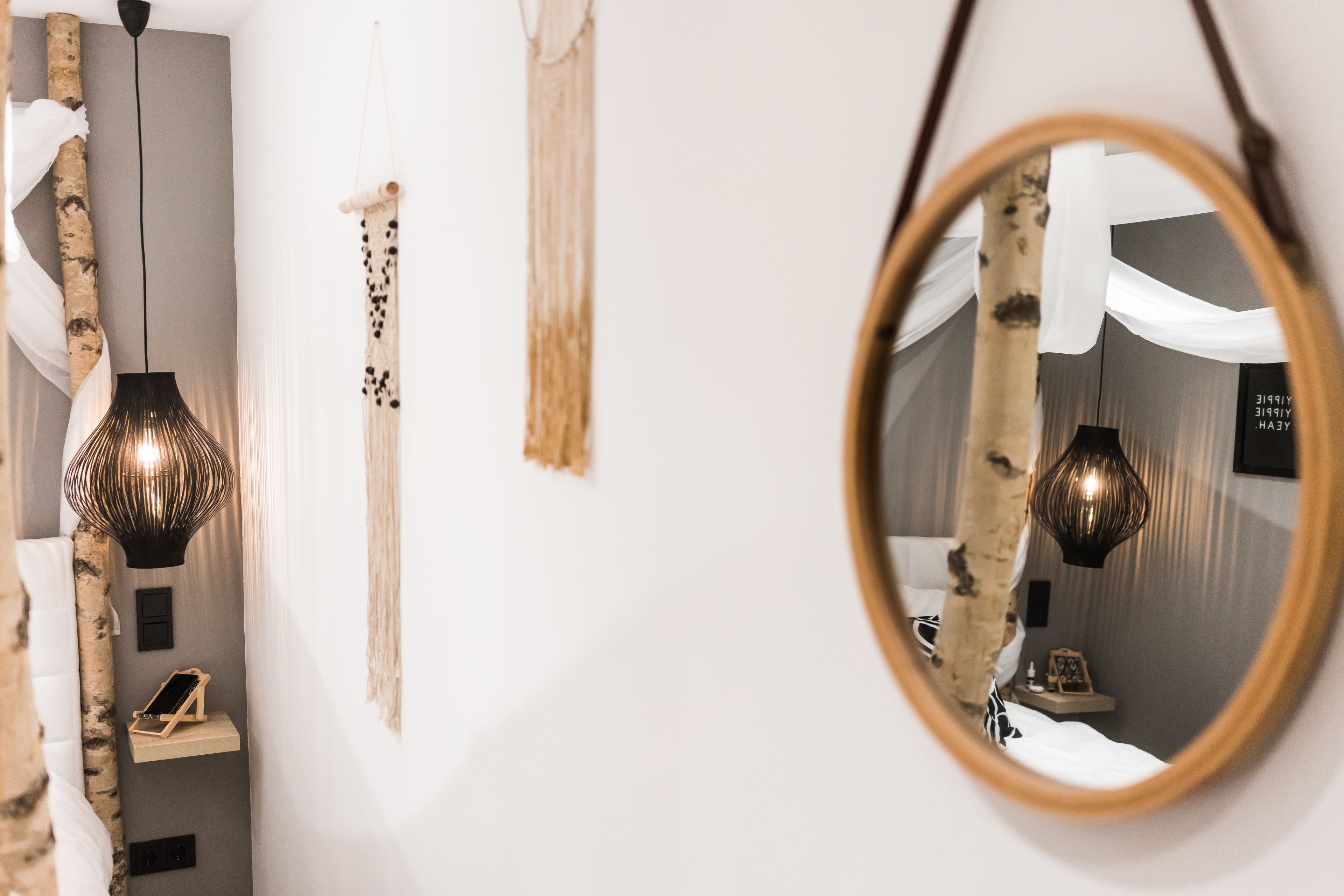 Een modern interieur met een grote spiegel die een kamer weerkaatst, met een bed, een hangende lamp en een wandplank met een klein voorwerp.