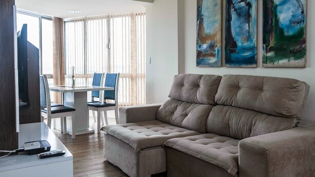 A living room with a beige sectional sofa, a dining table with chairs, and abstract paintings on the wall. - Habitat Ed. Diomício 1203 Urbano (Florianópolis)