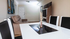 A dining area with a white table, black chairs, a beige sofa, and a TV mounted on the wall. - Habitat Ed. Diomício 1203 Urbano (Florianópolis)